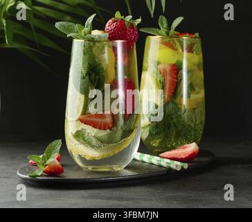 Ein erfrischendes Getränk im Glas mit Zitronenscheiben, Erdbeeren, frischen Minzblättern und Mineralwasser, serviert mit Limette und Minze auf einem schwarzen Teller Stockfoto