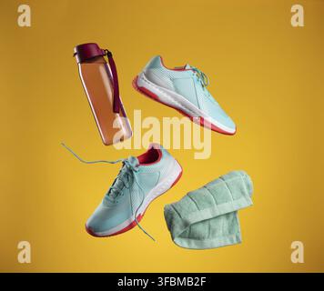 Rosa Plastikwasserflasche und grünes Handtuch sowie ein Paar blaue Textilsneaker mit Schnürsenkeln schweben auf einem gelben Hintergrund. Schuhe für Sport, Jogging Stockfoto