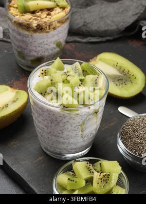 Zwei Gläser Chiasamenpudding, eines mit Kiwi-Würfel und das andere mit Müsli und Bananenchips, befinden sich auf einer dunklen Oberfläche mit Zutaten Stockfoto