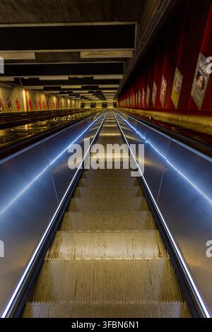 Blick auf eine lange, beleuchtete Rolltreppe in einer modernen U-Bahn-Station mit metallenen Seitenwänden und Werbeplakaten an der roten Wand in Stockholm, S Stockfoto