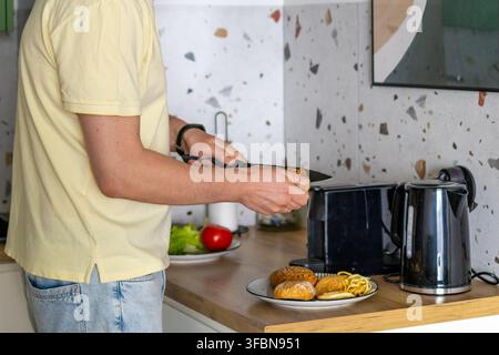 Mann schneidet frisches Brot auf Holzbrett für die Frühstücksvorbereitung in der modernen Küche zu Hause. Konzept der Mahlzeitvorbereitung und hausgemachten Frühstückskocherei Stockfoto
