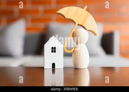 Versicherungskonzept für Haus und Leben mit hölzernem Regenschirm, der ein Miniaturhausmodell und eine Steckpuppenfigur auf einem reflektierenden Tisch mit Ziegelhintergrund bedeckt Stockfoto