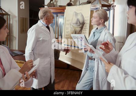 Senior weißer Professor erklärt eine Knöchelanatomie-Tafel diversen jungen medizinischen Studenten im Universitätsklassenzimmer. Bild geeignet für medizinische Bildungsmarketing Stockfoto