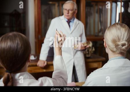 Junge kaukasische Medizinstudentinnen heben die Hände während der Vorlesung mit einem älteren männlichen Professor im Klassenzimmer. Bild geeignet für Marketing in Hochschulbildung und Medizinschulen Stockfoto