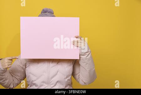 Frau in rosa Winterjacke und Hut hält rosafarbenes Blatt Papier auf gelbem Hintergrund. Saisonaler Verkauf. Platz für die Beschriftung Stockfoto