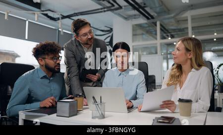 Vier multikulturelle Geschäftsleiter, die lachend Teamarbeit praktizieren, einen Laptop nutzen und Papiere, Dokumente, Statistiken, Ergebnisse, statistische rechtliche Daten überprüfen, multirassisch Stockfoto