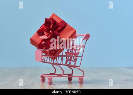 Ein in rotes Papier gewickeltes Geschenk in einem Miniatur-Einkaufswagen auf blauem Hintergrund Stockfoto