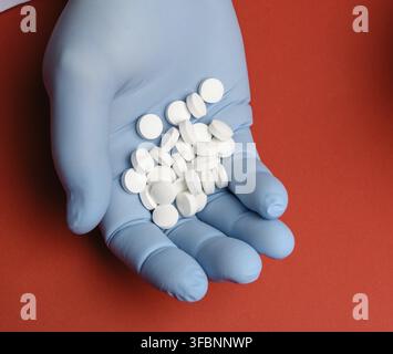Männliche Hand in einem blauen sterilen Gummihandschuh hält einen Stapel Pillen auf einem roten Hintergrund, Draufsicht Stockfoto