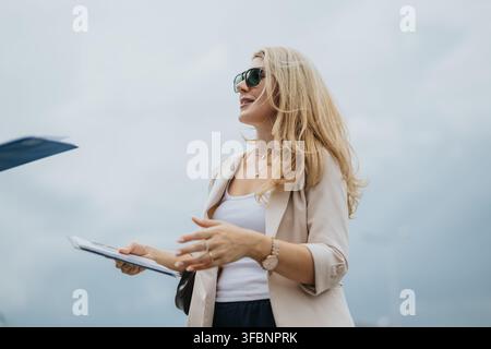 Selbstbewusste Geschäftsfrau mit Klemmbrett und Sonnenbrille steht im Freien in der Nähe eines Flugzeugs Stockfoto