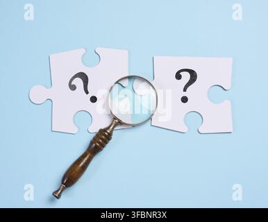 Zwei weiße Puzzleteile mit Fragezeichen unter einer Lupe auf blauem Hintergrund symbolisieren die Suche nach einer Lösung, Detektivuntersuchung Stockfoto