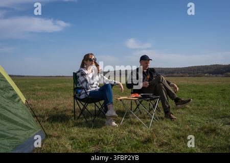 Paar genießt einen entspannten Picknickmoment, sitzt in Campingstühlen neben einem Zelt, trinkt zusammen Bier auf einer weiten grünen Wiese unter einem klaren blauen Himmel Stockfoto