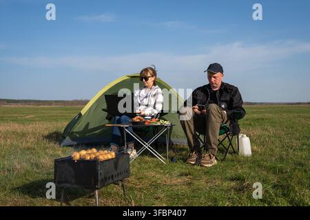 Paar arbeitet remote von einer sonnigen Wiese aus, Laptop und Telefon auf dem Schoß, grillt über einem Grill neben ihrem Zelt, genießt die Freiheit als Digital Nomads und o Stockfoto