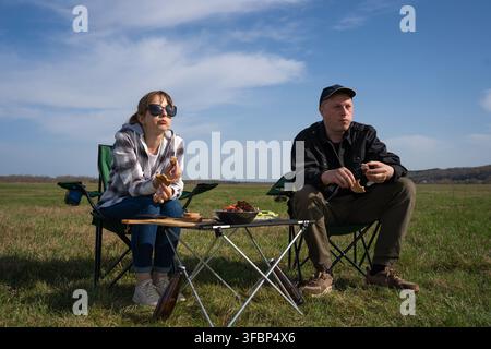 Paar sitzt auf Campingstühlen, isst Barbecue und feiert ein Picknickmittagessen in einem offenen Feld und schafft einen Moment der Entspannung und Gemeinschaft Stockfoto