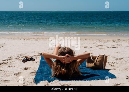 Frau liegt auf einem blauen Handtuch mit den Händen hinter dem Kopf auf leerem weißem Sand mit Blick auf das offene Meer. Solo-Reise und unabhängiger Sommerurlaub. Stockfoto