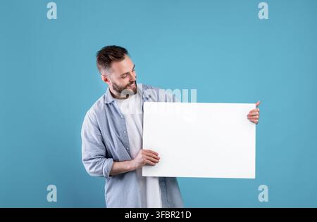 Glücklich lächelnder Mann, der weiße leere Plakette auf blauem Studiohintergrund anzeigt, mockup für Logo oder Design Stockfoto