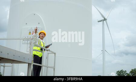 Professionelle asiatische Ingenieurin trägt Sicherheitshelm und reflektierende Weste, hält ein Tablet und inspiziert einen Windturbinen-Generator an einem erneuerbaren Ort Stockfoto