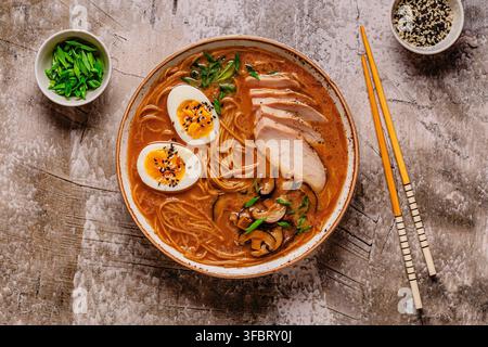 Köstliche Schüssel Ramen mit Hühnchen, Eiern, Pilzen und grünen Zwiebeln, serviert auf einem strukturierten Tisch Stockfoto