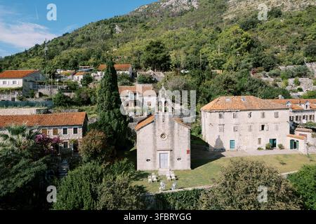 Ruhiges Steindorf unter steilen grünen Bergen, mit Kapelle, Terracotta-Dächern, Zypressen und sonnenbeschienenen mediterranen Häusern, die sich über das Dorf verteilen Stockfoto