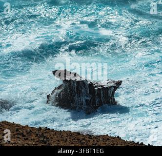 dunkler Vulkanfelsen, umgeben von mächtigen türkisfarbenen Meereswellen und weißem Schaum entlang einer zerklüfteten Küste. Stockfoto