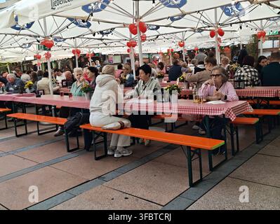 Pforzheim Deutschland 23. August 2026 Menschenmenge, die an langen Holztischen mit rot karierten Tischdecken unter großen weißen Sonnenschirmen im Freien sitzt Stockfoto