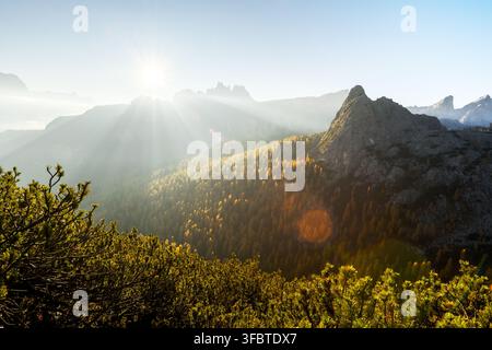 Sonnenstrahlen brechen durch den Bergnebel über Herbstwäldern in den Dolomiten. Atmosphärische Alpenlandschaft mit goldenen Lärchen und geschichteten Bergkämmen Stockfoto