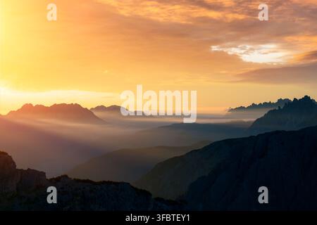 Goldenes Sonnenlicht breitet sich über Bergtäler und entfernte Kämme in den Dolomiten aus. Atmosphärische Landschaft mit Sonnenstrahlen, Dunst und geschichteten alpinen Silhouetten Stockfoto