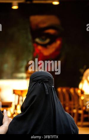 Eine Person in schwarzer Kleidung steht vor einem großen Gemälde, das ein Gesicht mit einem Auge und roten Markierungen zeigt. Stockfoto