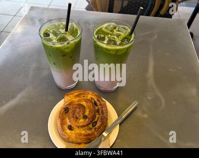 Zwei Vanille-, Blaubeer- und Erdbeer-Iced Matcha mit schwarzen Strohhalmen und ein Pain au Raisin auf einem Stahltisch in einem Café Stockfoto