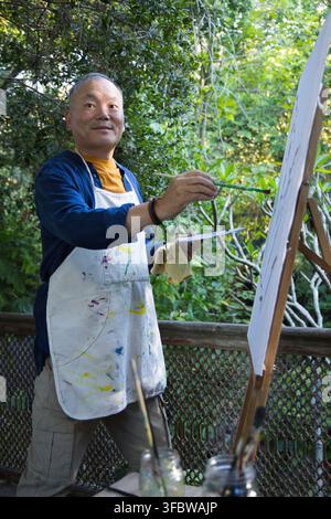 Asiatischer Senior mit einem mit Farbe befleckten Schürzen, der auf dem Deck mit Staffelei, Pinselgläsern und Palette malt Stockfoto