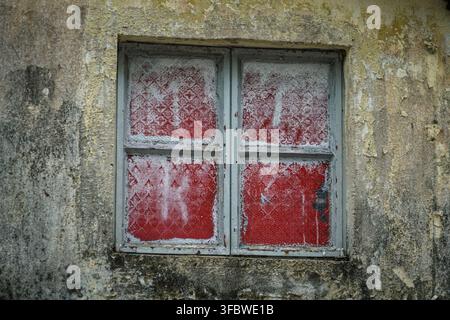 Ein stark beschädigter hellgrauer Fensterrahmen mit vier Scheiben ist in eine strukturierte, abblätternde, gelb-braune Betonwand eingebettet. Die Fensterscheiben sind cove Stockfoto