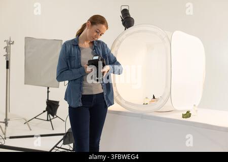 Frau in ihren 20ern überprüft DSLR-Einstellungen, während sie an einem Tisch in der Nähe eines Lichtzeltes mit Flaschen lehnt Stockfoto