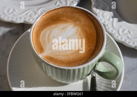 Ein frisch gebrühter Cappuccino mit zarter Blatt-Latte-Art, serviert in einer gerippten hellgrünen Tasse mit Untertasse. Stockfoto