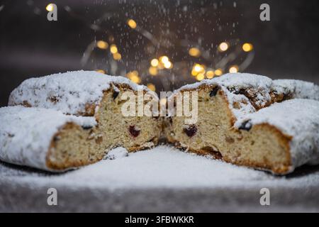 Eine Nahaufnahme eines traditionellen Weihnachtsstollens mit Früchten, der mit Puderzucker bestäubt wird, vor einem dunklen Hintergrund mit festlichen Bokeh-Lichtern. Stockfoto