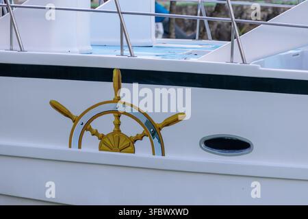 Nahaufnahme eines weißen Yachtrumpfes mit einem dekorativen goldenen Schiffsrad, einem ovalen Bullauge und Metallgeländern an einem ruhigen Marinetag. Stockfoto