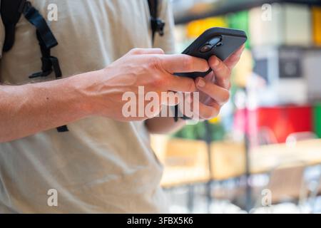 Nahaufnahme von männlichen Händen, die ein schwarzes Smartphone mit einem Rucksack halten und darauf tippen, online verbunden oder remote arbeiten in einem hellen unscharfen urbanen Hintergrund Stockfoto