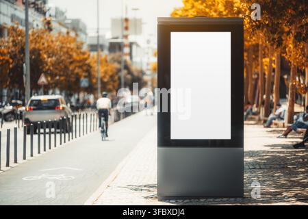 Leere vertikale Werbetafel steht neben einem sonnigen Stadtfahrradweg, mit einer entfernten Fahrradfahrer-Silhouette links zwischen Verkehr, Bäumen und warmem u Stockfoto