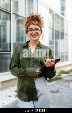 Eine Frau lächelt vor einem Glasgebäude, während sie ein Klemmbrett hält. Sie trägt eine Brille und ein grünes Hemd. Stockfoto