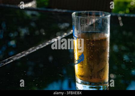 Ein klares Glas gefüllt mit einem bernsteinfarbenen kohlensäurehaltigen Getränk, wahrscheinlich Bier, das auf einem dunklen Außentisch steht. Stockfoto