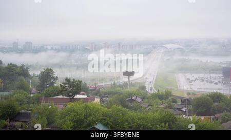 Luftaufnahme einer Stadt Irkutsk, Landschaft bedeckt von dichtem Morgennebel, Russland. Mehrspurige Autobahn und Brücke, die zu einer modernen Skyline führen, Nebel über Stockfoto