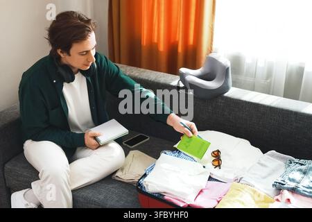 Mann packt Gepäck für eine Reise. Er sitzt auf einem Sofa, überprüft seinen Planungsnotizblock und legt seinen Pass in den Koffer für den Urlaub. Hohe q Stockfoto