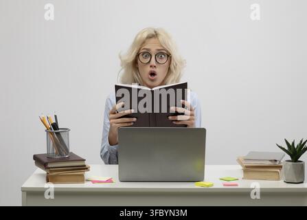 Überraschte junge Frau hält ein Buch im Büro und schaut schockiert beim Lernen Stockfoto