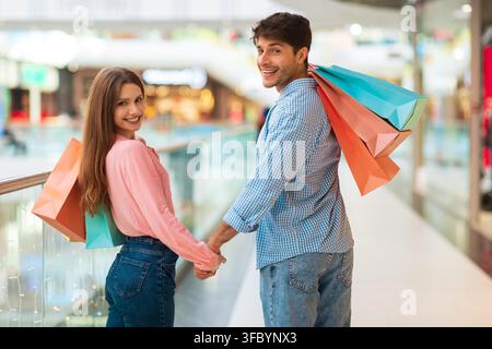Glückliches junges Paar, das Händchen hält und zusammen Einkaufstüten in der Einkaufs mall trägt Stockfoto
