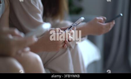 Nahaufnahme von jungen Frauen, die drinnen im weichen natürlichen Licht auf Smartphones scrollen, lässige beige Kleidung, moderner Lebensstil-Moment des Digitalen Stockfoto