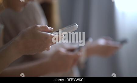 Freunde, die zu Hause in ihre Handys vertieft sind, Hände halten Geräte, konzentriert auf Bildschirme, entspannte Innenraumatmosphäre mit sanftem Fensterlicht und Stockfoto