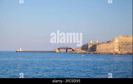 Eine Steinfortress und ein Leuchtturm befinden sich entlang eines langen Piers und eines Molens, während ein kleines Boot auf ruhigem blauem Wasser unter einem klaren Abendhimmel treibt. Valletta, Malta Stockfoto
