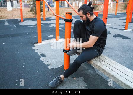 Mann in schwarzer Sportkleidung sitzt auf Holzbank im Outdoor-Fitnesspark, bindet Laufschuh neben leuchtend orangefarbenen Fitnessstangen und abgenutzten Pflastersteinen Stockfoto