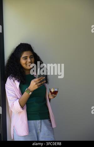 Indische Frau lehnt am Türrahmen, lächelt, überprüft das Telefon, hält eine Teetasse, eine Smartwatch, Kopierraum Stockfoto