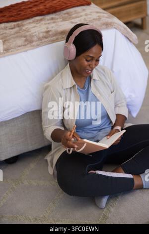 Frau sitzt auf einem Teppich im Schlafzimmer, trägt rosa Kopfhörer und hält ein rosa Notizbuch und einen roségoldenen Stift Stockfoto