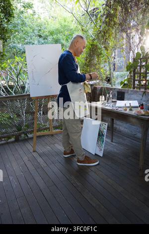 Seniorer asiatischer Mann steht auf der Terrasse in einer gefärbten Schürze und hält eine Palette, Pinsel zum Vorbereiten von Farben Stockfoto