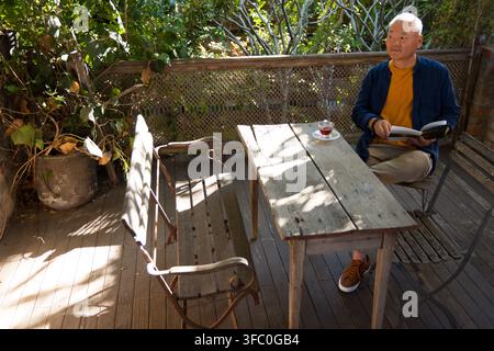 Asiatischer Senior sitzt auf dem Steuerdeck und liest ein Buch mit einer Teetasse, trägt ein Senfhemd, Kopierraum Stockfoto